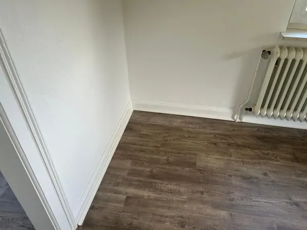 Leere Zimmerecke mit weißer Wand, Sockelleiste und Heizkörper am Fenster