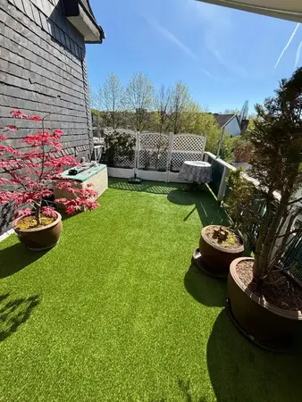 Sonniger Balkon mit Kunstrasen, Pflanzkübeln, Sichtschutz und Tisch.